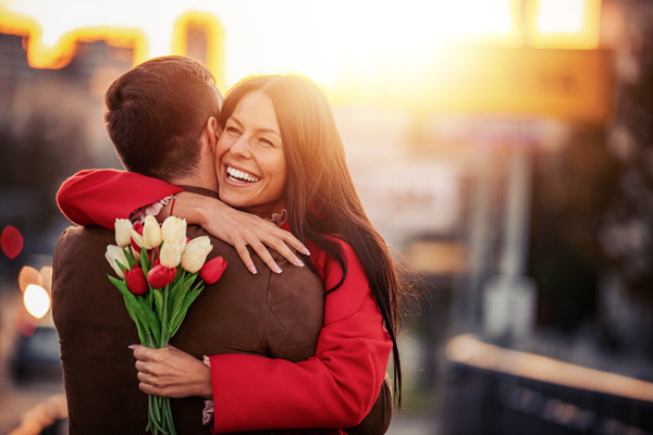 Foto: Casal de namorados se abraçando. A mulher sorri segurando um buquê de tulipas vermelhas e brancas.